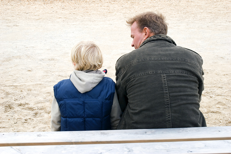 A father and sun sitting together on a beach in winter coats with their backs to the camera.