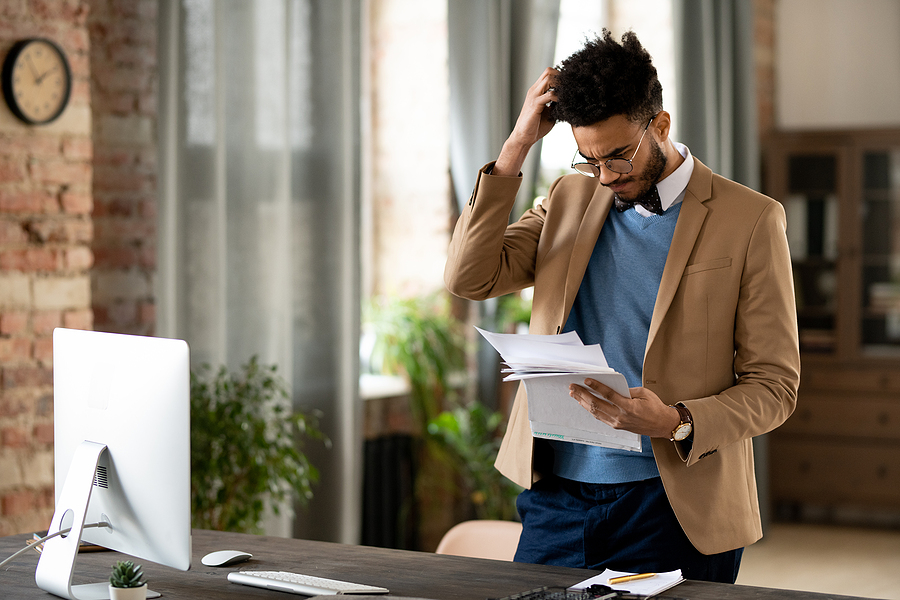 Confused young mixed race man in glasses and jacket standing at desk with computer and scratching head while reading papers