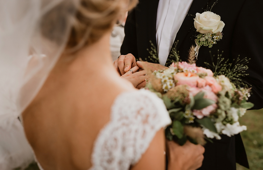 The groom is putting the wedding ring on the bride. Image has been taken over the shoulder of a bride without faces of the couple. Focus on hands of newlyweds, foreground and background out of focus.