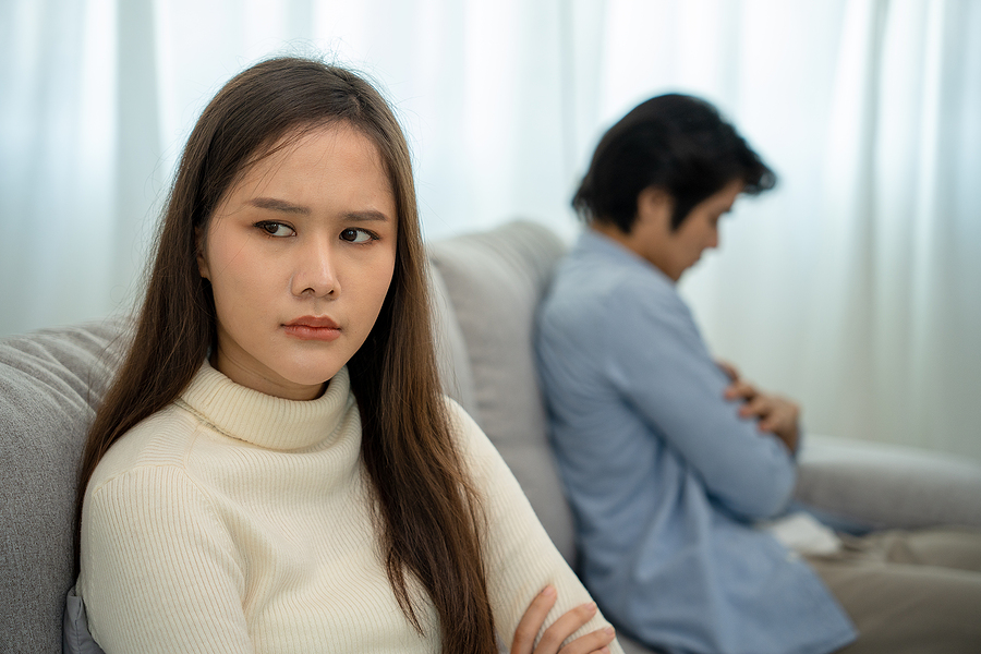 Couple sitting apart from each other on a couch after a quarrel - wife looking towards the camera displeased, husband facing the other way