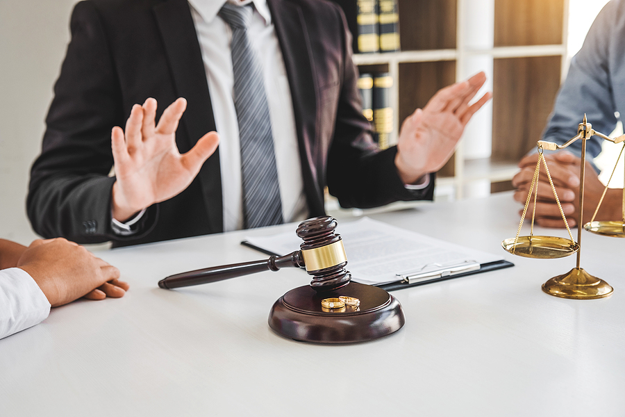 Couple sitting across from each other at a lawyers desk, lawyer sitting between them settling the dispute. Gavel, paperwork, and scale sits on the desk.