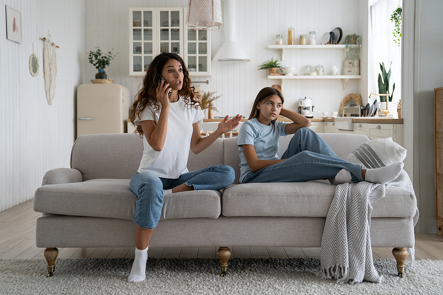 Young mother stressed while talking on the phone about her divorce and child support issues. Child sitting on the couch next to her looking annoyed.