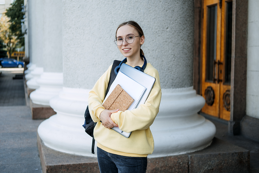 Young girl standing in front of a college campus building wearing a yellow sweater, carrying a backpack on her shoulder, with notebooks in her arms.