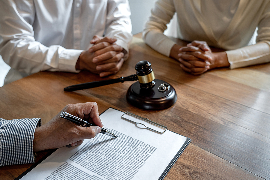 Photo of a couple sitting at a divorce lawyer's desk while divorce lawyer sits across from them reading from clip board, a gavel sits on the table between the three of them