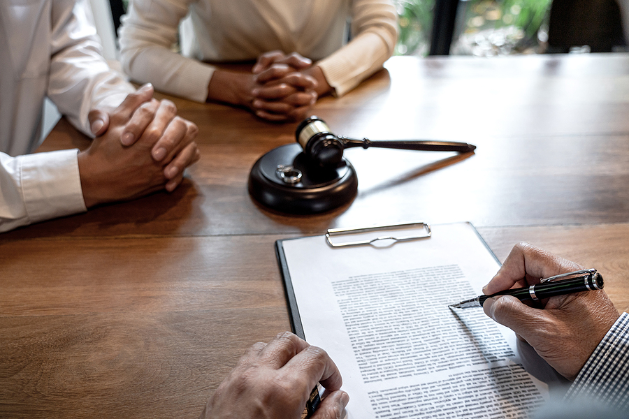 Couple sitting across from divorce lawyer with their hands folded, lawyer writing on paperwork on a clipboard with a pen