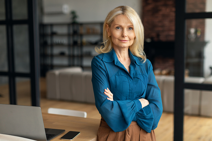 Portrait of confident smiling mature businesswoman sitting leaning on desk posing with folded arms at home office
