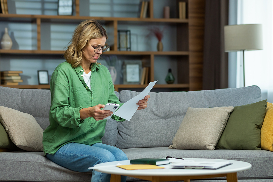Senior beautiful woman in glasses sitting on sofa at home. Worriedly holding the phone and documents in her hands. Problem with accounts, credit, payment.