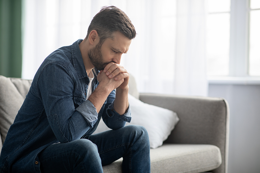 Sad bearded middle-aged man sitting on couch at home, leaning on hands and looking down, upset man having problems, side view, copy space. Loneliness, depression